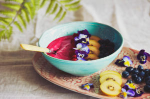 High angle close up of bowl with pink fruit puree with blueberries and slices of yellow kiwis, decorated with pansy petals. High angle close up of bowl with pink fruit puree with blueberries and slices of yellow kiwis, decorated with pansy petals.